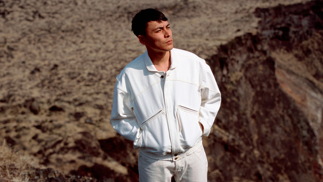 Image shows man wearing a white denim jacket, white pants, squinting his eyes as he looks in the direction of the sun. The man stands on the edge of a volcanic crater. 